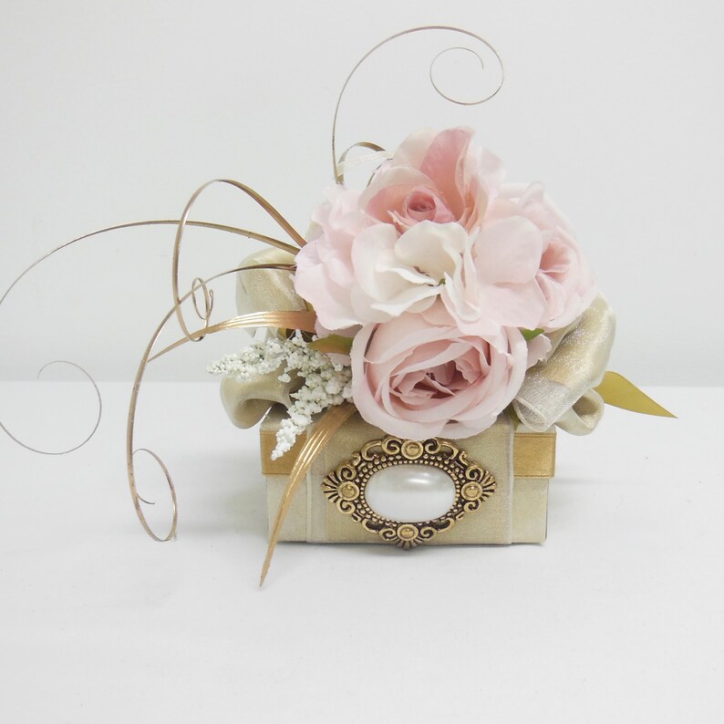 K&ouml;nnte beinhalten: Eine kleine, quadratische, beige Box mit goldener Verzierung und einer dekorativen goldenen Brosche mit einer Perle in der Mitte. Die Box ist mit einem Strau&szlig; rosa Rosen und wei&szlig;en Blumen verziert.