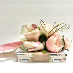 K&ouml;nnte beinhalten: Eine dekorative Geschenkbox mit floralem Muster, verziert mit rosa und gr&uuml;nen B&auml;ndern und k&uuml;nstlichen rosa Rosen. Die rechteckige Box hat ein passendes florales Band.