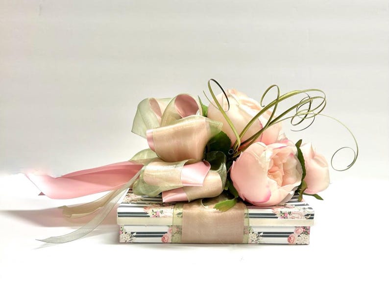 K&ouml;nnte beinhalten: Eine dekorative Geschenkbox mit floralem Muster, verziert mit rosa und gr&uuml;nen B&auml;ndern und k&uuml;nstlichen rosa Rosen. Die rechteckige Box hat ein passendes florales Band.