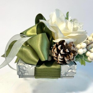 K&ouml;nnte beinhalten: Eine kleine, wei&szlig;e Geschenkbox mit einem gr&uuml;nen Satinband und Schleife. Die Box ist mit einem Blumenmuster verziert und mit einer wei&szlig;en Rose, einem Tannenzapfen und wei&szlig;en Beeren gekr&ouml;nt.