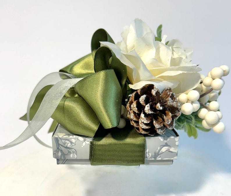 K&ouml;nnte beinhalten: Eine kleine, wei&szlig;e Geschenkbox mit einem gr&uuml;nen Satinband und Schleife. Die Box ist mit einem Blumenmuster verziert und mit einer wei&szlig;en Rose, einem Tannenzapfen und wei&szlig;en Beeren gekr&ouml;nt.