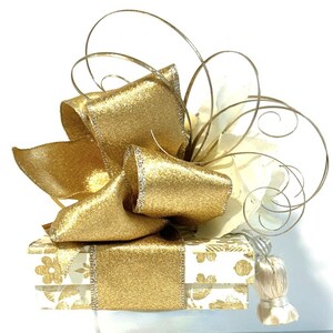 May include: A white gift box with a gold floral pattern is topped with a large gold bow and a white and gold decorative accent.