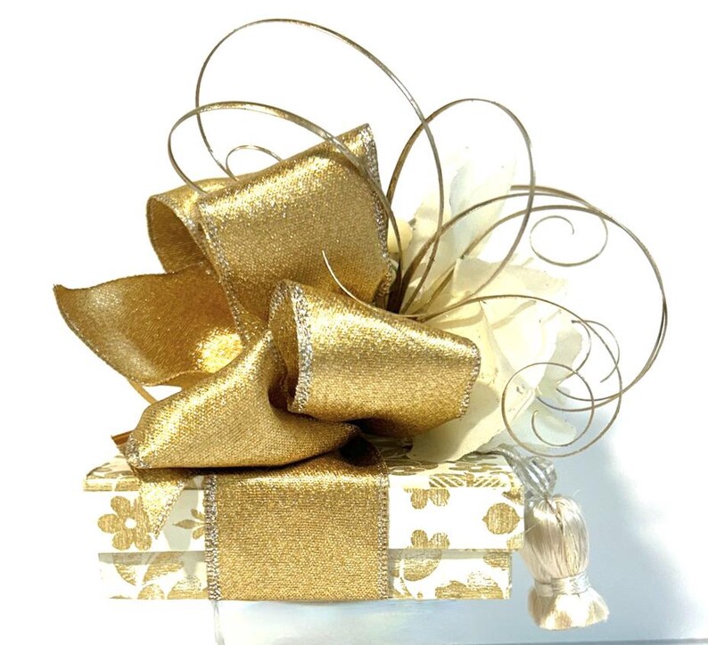 May include: A white gift box with a gold floral pattern is topped with a large gold bow and a white and gold decorative accent.