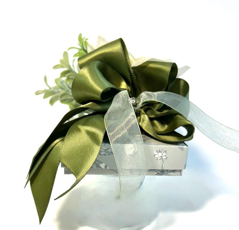 K&ouml;nnte beinhalten: Eine kleine Geschenkbox mit einem gr&uuml;nen Satinband und Schleife. Die Box ist mit einem Blumenmuster verziert und hat einen wei&szlig;en Hintergrund.