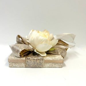 Puede incluir: Una caja de regalo envuelta en papel estampado beige y marr&oacute;n con una flor blanca en la parte superior. La caja est&aacute; atada con una cinta beige con ribete dorado.