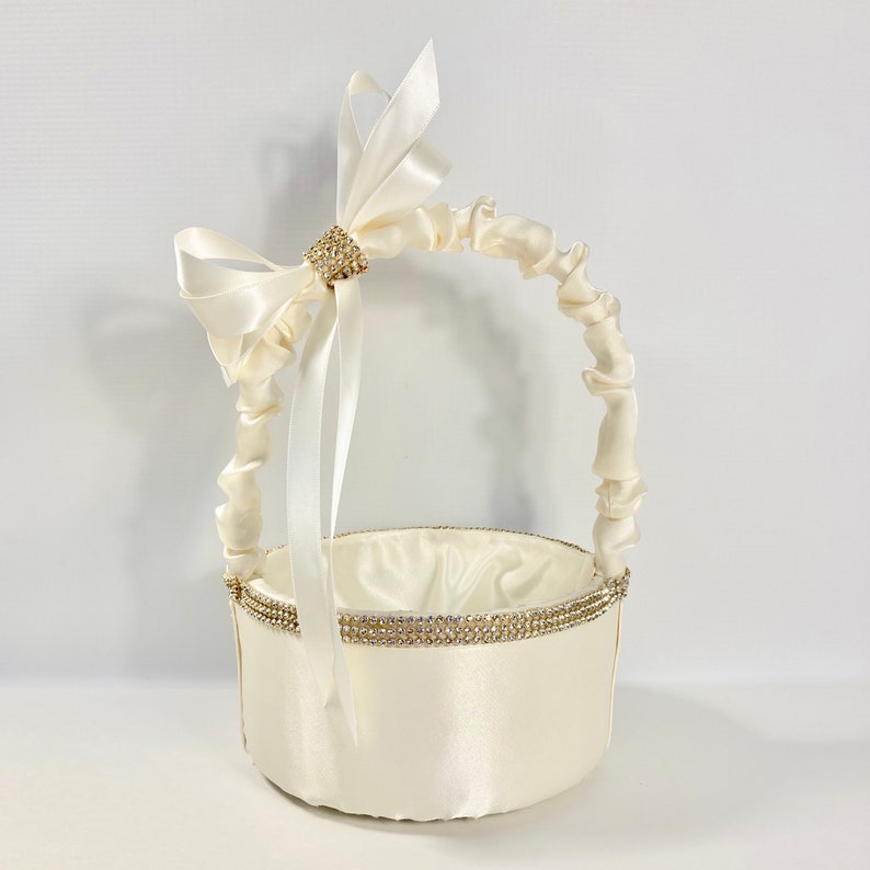 May include: A white satin flower girl basket with a white satin bow and a rhinestone embellishment. The basket has a ruffled handle.