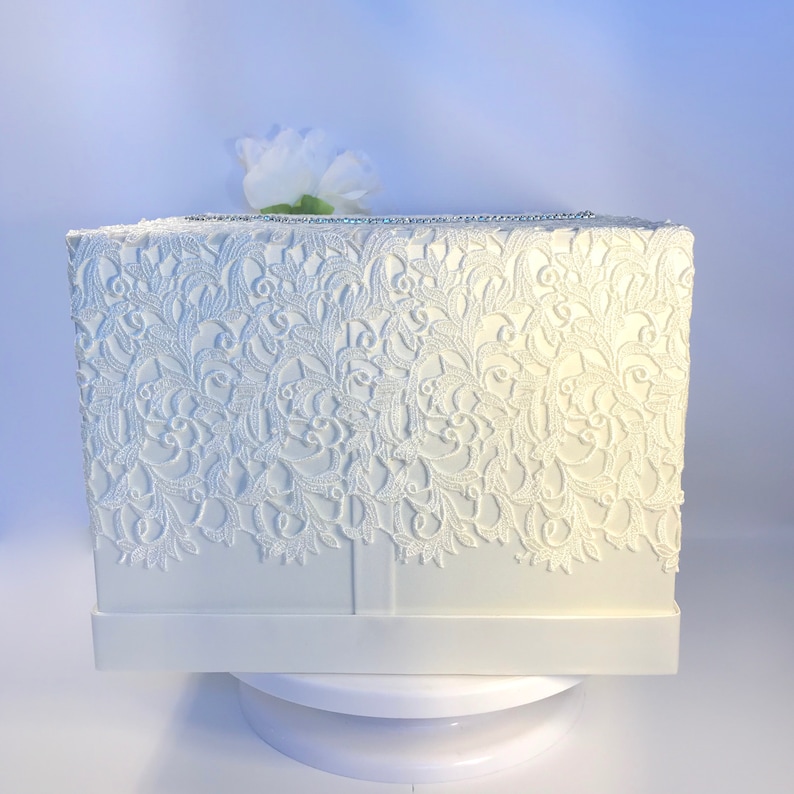 May include: A white rectangular box with detailed, lace-like patterns on the front and sides. A small white flower rests on top. The box has a decorative trim along the top edge. The box is on a white platform.