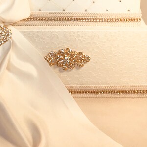 May include: Ivory-colored decorative box with a textured surface, adorned with gold trim and sparkling embellishments. A flowing ivory fabric is draped over the box, secured by a jeweled brooch. The box features a diamond pattern and additional gold accents.