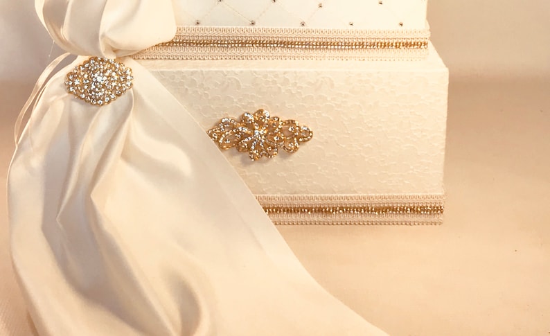 May include: Ivory-colored decorative box with a textured surface, adorned with gold trim and sparkling embellishments. A flowing ivory fabric is draped over the box, secured by a jeweled brooch. The box features a diamond pattern and additional gold accents.