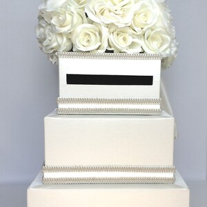 May include: A white wedding card box with three tiers. The top tier has a slot for cards and is decorated with a white rose bouquet. The box is trimmed with a silver beaded ribbon.