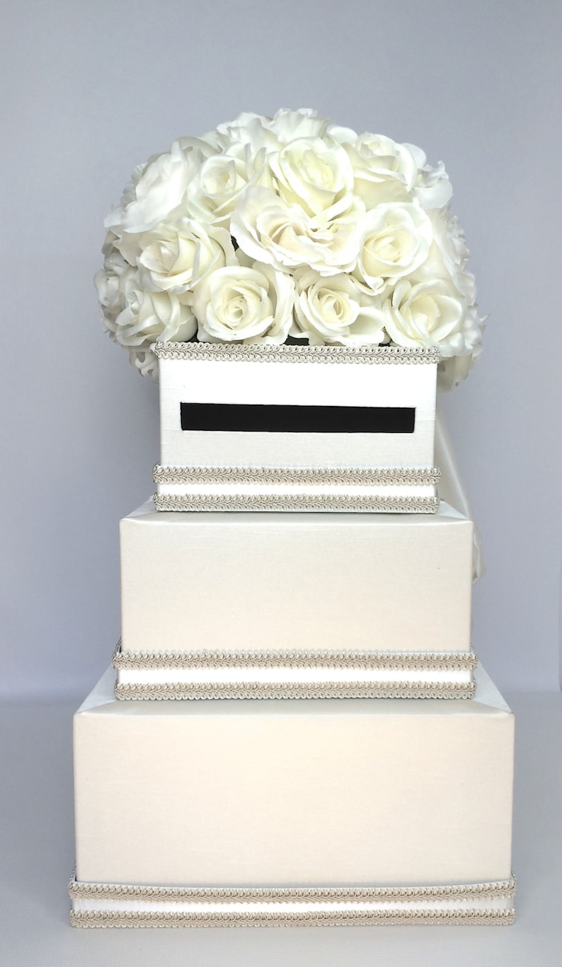 May include: A white wedding card box with three tiers. The top tier has a slot for cards and is decorated with a white rose bouquet. The box is trimmed with a silver beaded ribbon.