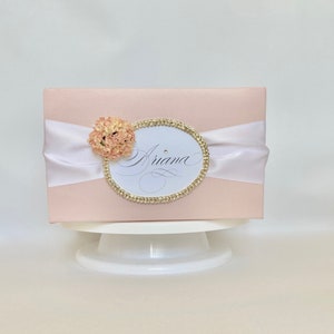 May include: A light pink gift box with a white satin ribbon and a decorative oval. The oval is trimmed with gold rhinestones and features the name "Ariana" in elegant script. A small peach-colored flower is attached to the left side of the oval.