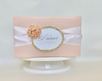 May include: A pale pink gift box with a white satin ribbon and an oval embellishment. The oval is edged with gold rhinestones and displays the name "Ariana" in cursive. A small peach-toned flower is attached to the left side of the oval.