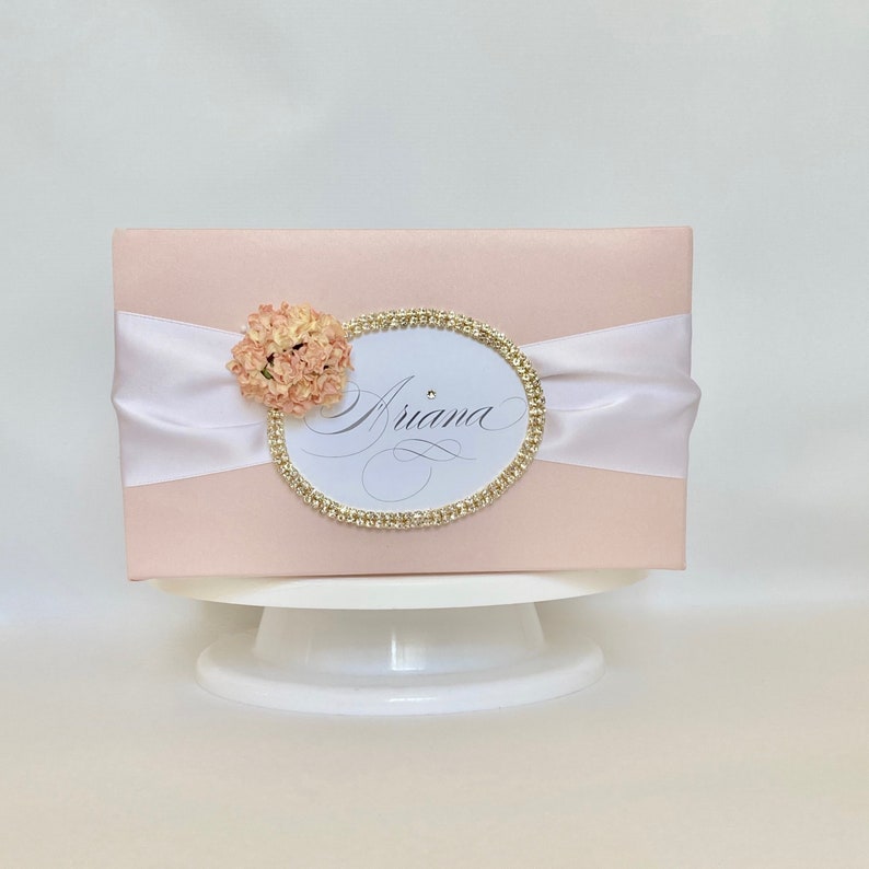 May include: A light pink gift box with a white satin ribbon and a decorative oval. The oval is trimmed with gold rhinestones and features the name "Ariana" in elegant script. A small peach-colored flower is attached to the left side of the oval.