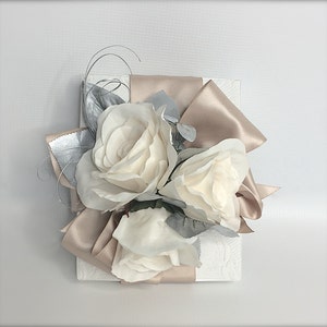 May include: A white gift box with a satin ribbon bow in a light brown color. The bow is decorated with three white roses and silver leaves.