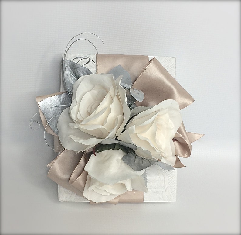 May include: A white gift box with a satin ribbon bow in a light brown color. The bow is decorated with three white roses and silver leaves.