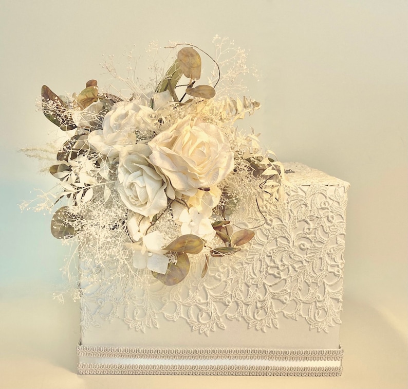 May include: White lace wedding card box with a bouquet of white flowers and greenery on top.