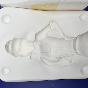 May include: A white ceramic slip casting mould featuring a recessed relief of a figure with a bun hairstyle, wearing a collared garment with ruffled sleeves. The rectangular mould has four circular indentations at the corners for alignment.