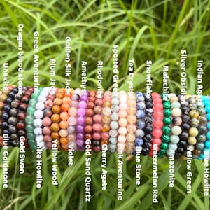 Peut inclure: Collection de bracelets de perles de différentes couleurs et pierres précieuses, dont l'améthyste, le jade et l'obsidienne. Les bracelets sont présentés sur une surface rose devant de l'herbe verte. Chaque bracelet est étiqueté avec le nom de sa pierre.