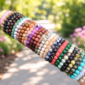 May include: A collection of numerous beaded bracelets featuring a wide variety of natural stones in diverse colors including blue, green, pink, purple, black, and white. The round beads are strung together in a long, stacked row on a cylindrical display.