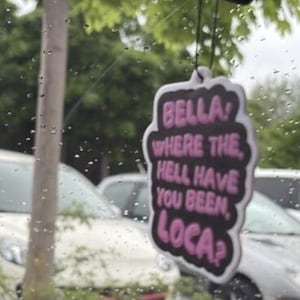 May include: A car air freshener with a black background and a white border. The text "BELLA! WHERE THE HELL HAVE YOU BEEN, LOCA?" is written in pink. The air freshener is hanging in front of a window with a blurred outdoor scene.
