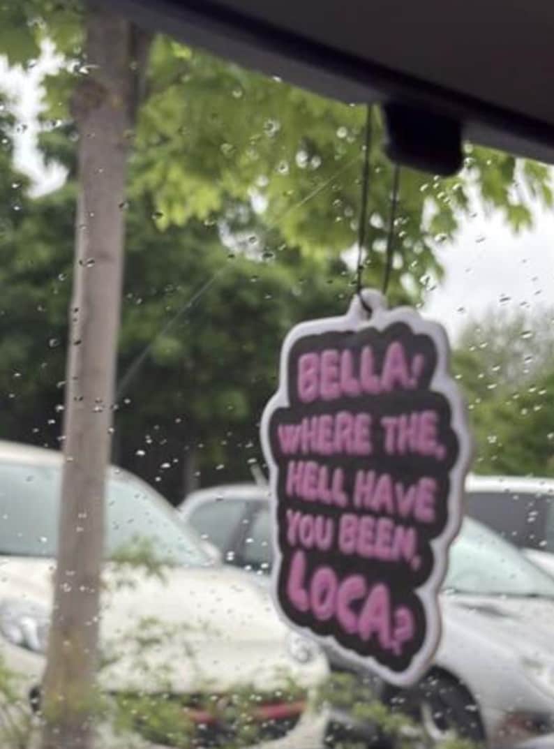 May include: A car air freshener with a black background and a white border. The text "BELLA! WHERE THE HELL HAVE YOU BEEN, LOCA?" is written in pink. The air freshener is hanging in front of a window with a blurred outdoor scene.