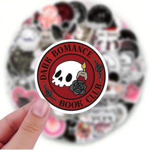 May include: A circular sticker featuring the text "DARK ROMANCE BOOK CLUB" around a white skull, a lit candle, and a grey rose on a red backdrop. The sticker is held by a hand, with other stickers blurred in the background.