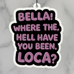 Op de afbeelding: Een witte luchtverfrisser met een zwarte rand en de tekst "BELLA! WHERE THE, HELL HAVE YOU BEEN, LOCA?" in paars. De luchtverfrisser hangt aan een zwart touwtje.