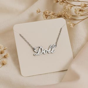 Könnte beinhalten: Eine silberfarbene Halskette mit dem Wort "Doll" in Schreibschrift. Die Halskette wird auf einer weißen Karte präsentiert, vor einem weichen, neutralen Hintergrund mit Trockenblumen.