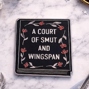 May include: Black enamel pin with the text "A Court of Smut and Wingspan" in white. The pin is square with a floral border of red and silver accents. The pin is on a marble surface.