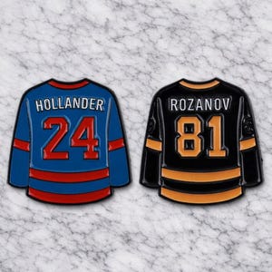 May include: Two hockey jersey enamel pins. One is blue with red stripes, the name "HOLLANDER" and the number "24" in red. The other is black with orange stripes, the name "ROZANOV" and the number "81" in orange.