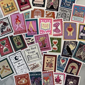 May include: A collection of colorful, vintage-style stickers with various romantic themes. Designs include illustrations of books, hearts, flames, and text such as "Vampire Romance" and "Enemies to Lovers". The stickers are arranged on a light surface.