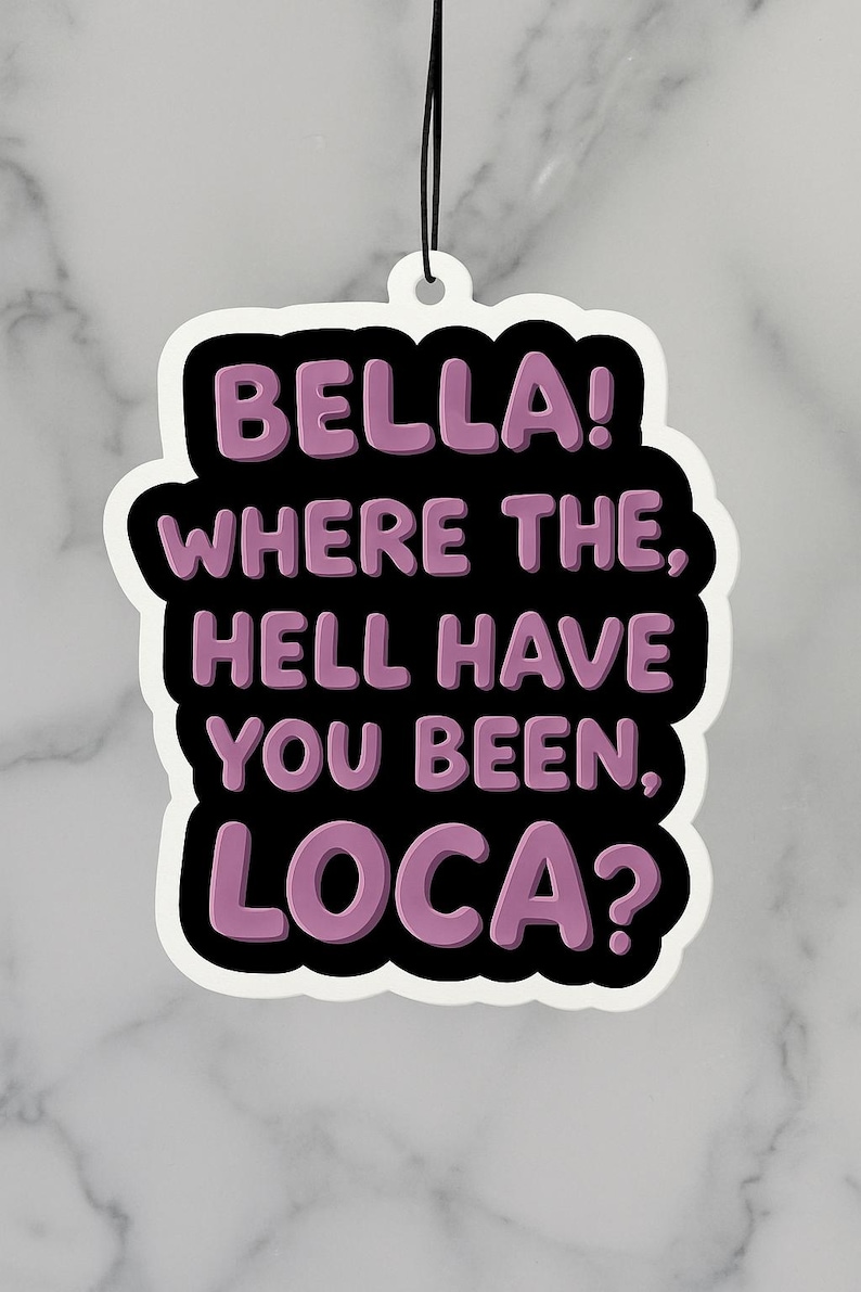 Op de afbeelding: Een witte luchtverfrisser met een zwarte rand en de tekst "BELLA! WHERE THE, HELL HAVE YOU BEEN, LOCA?" in paars. De luchtverfrisser hangt aan een zwart touwtje.