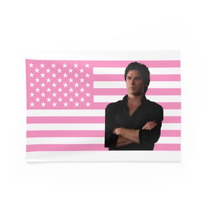 May include: A rectangular wall tapestry featuring a pink and white American flag design. A person in a black shirt is positioned to the right of the flag, with arms crossed. The tapestry is displayed against a white background.