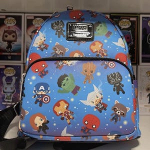 Könnte beinhalten: Blauer Mini-Rucksack im Marvel-Design mit verschiedenen Superhelden-Charakteren. Der Rucksack hat eine Reißverschlusstasche vorne und verstellbare Träger. Das Marvel-Logo ist auf einem schwarzen Patch zu sehen. Im Hintergrund befinden sich Funko Pop-Boxen.