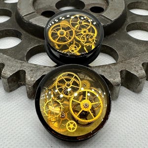 May include: Two black ear gauges with a clear, domed top showcasing intricate golden clockwork gears. The gears are of varying sizes and are set against a light yellow background. The gauges are displayed on a metal gear.