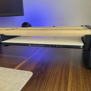 May include: A desk accessory featuring a series of light-coloured wooden dowels arranged horizontally above a flat, white rectangular surface. The structure is supported by black side panels, and the text AXIONLAYER is printed on the front edge.