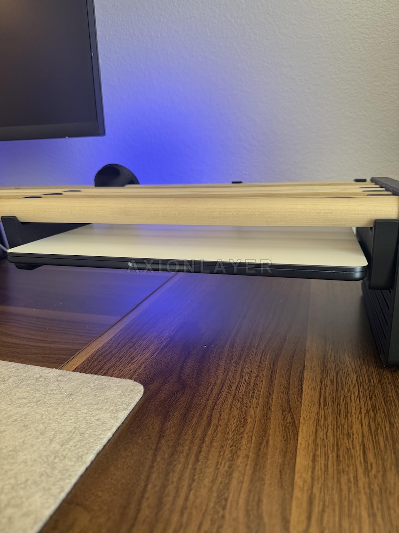 May include: A desk accessory featuring a series of light-coloured wooden dowels arranged horizontally above a flat, white rectangular surface. The structure is supported by black side panels, and the text AXIONLAYER is printed on the front edge.