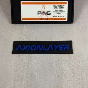 May include: Black rectangular label with the word "AXIONLAYER" in blue lettering. Above it is a black rectangular case with a white and orange label that reads "PING" and "KARSTEN MFG. CORP." Made in U.S.A.