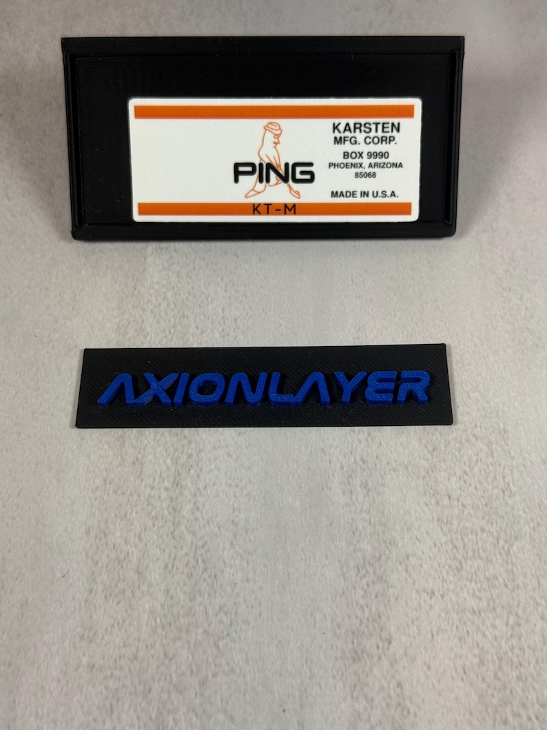 May include: Black rectangular label with the word "AXIONLAYER" in blue lettering. Above it is a black rectangular case with a white and orange label that reads "PING" and "KARSTEN MFG. CORP." Made in U.S.A.