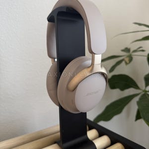 May include: Beige over-ear wireless headphones with gold-tone accents and the Bose logo on the ear cups, mounted on a vertical black stand. The headphones feature a padded headband and soft ear cushions, with the text AXIONLAYER visible.