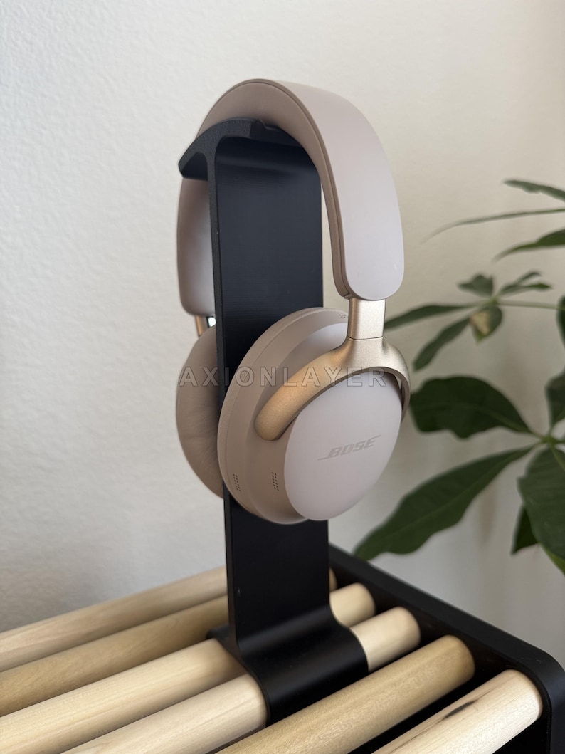 May include: Beige over-ear wireless headphones with gold-tone accents and the Bose logo on the ear cups, mounted on a vertical black stand. The headphones feature a padded headband and soft ear cushions, with the text AXIONLAYER visible.