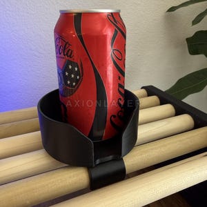 May include: A black plastic cup holder with a clip-on attachment, currently holding a red Coca-Cola soda can. The holder features a circular design with a cutout section and is secured onto a slatted wooden surface. The text AXIONLAYER is visible.