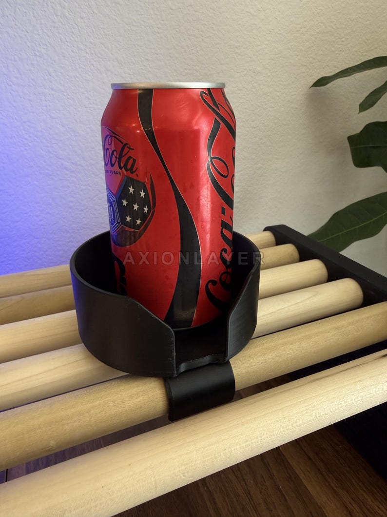 May include: A black plastic cup holder with a clip-on attachment, currently holding a red Coca-Cola soda can. The holder features a circular design with a cutout section and is secured onto a slatted wooden surface. The text AXIONLAYER is visible.