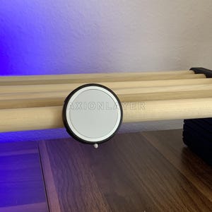 May include: A circular white magnetic charging puck with a black textured rim, featuring the text AXIONLAYER printed in white across the centre. The device is mounted onto a light-coloured wooden dowel rod structure.