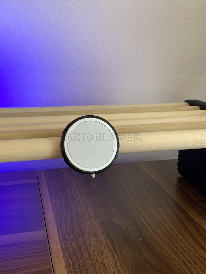 May include: A circular white magnetic charging puck with a black textured rim, featuring the text AXIONLAYER printed in white across the centre. The device is mounted onto a light-coloured wooden dowel rod structure.