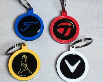 May include: Four round keychains with silver key rings. The keychains have a black center with a logo or letter in the center. The keychains are blue, red, yellow, and white.