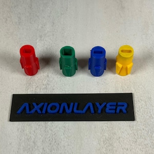 Könnte beinhalten: Vier farbenfrohe, 3D-gedruckte Düsen in Rot, Grün, Blau und Gelb. Jede Düse hat eine einzigartige Form. Darunter ein schwarzes Rechteck mit dem blauen Text "AXIONLAYER".