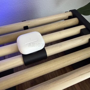 May include: A white wireless earbud charging case with the text AXIONLAYER printed on the front, mounted on a black clip attached to a light-coloured wooden slat rack. The case features a small circular indicator light on its smooth surface.