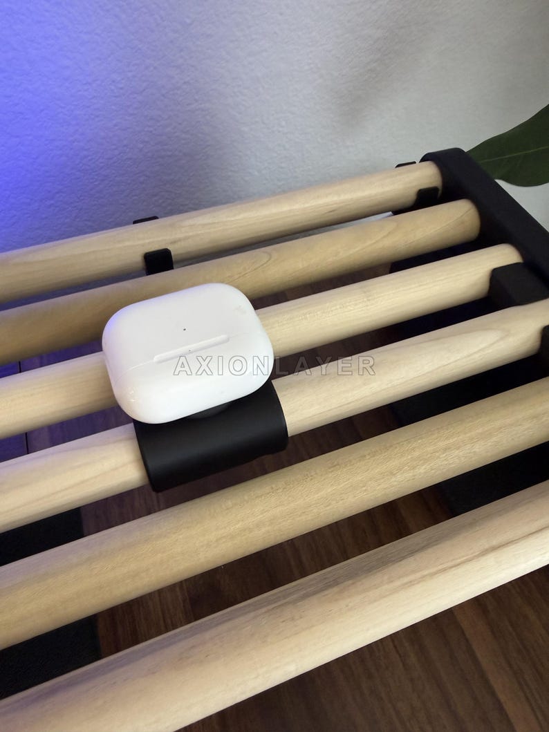 May include: A white wireless earbud charging case with the text AXIONLAYER printed on the front, mounted on a black clip attached to a light-coloured wooden slat rack. The case features a small circular indicator light on its smooth surface.